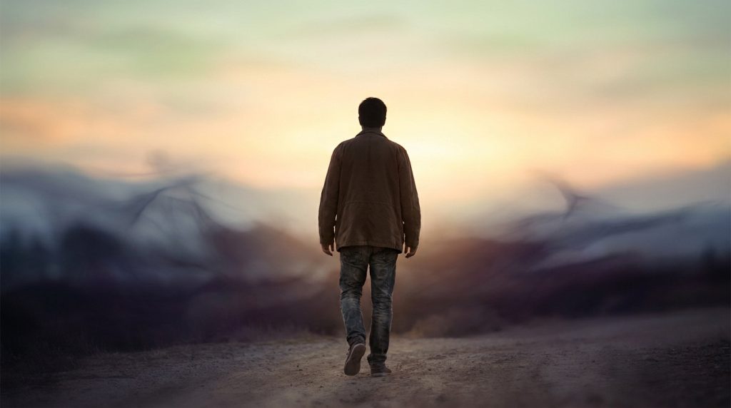 Un homme marche seul vers l'horizon au coucher du soleil, entouré de silhouettes vaporeuses symbolisant le passé.